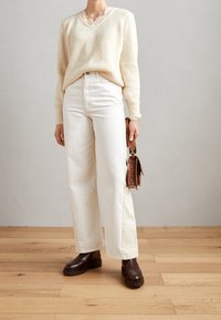 Crème gebreide trui met een diepe V-hals, gecombineerd met hoge witte wijde broek en bruine stevige enkellaarzen, terwijl er een bruine tas wordt vastgehouden.