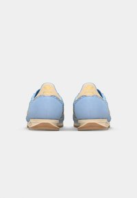 Zapatillas deportivas de ante azul con detalles en dorado, texturizados y una media suela beige, vistas desde atrás sobre un fondo blanco.