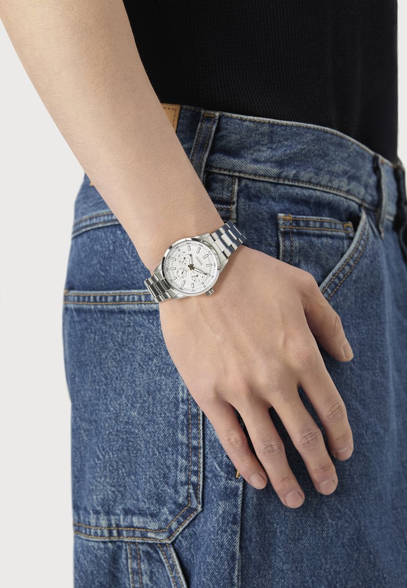 Sølvsømløs rustfritt stål klokke med rund klokkehus, hvit strukturert skive og sølvfargede timemarkører, plassert på en denim bakgrunn.