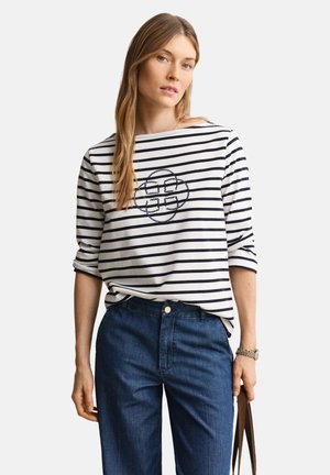 Mujer con camiseta de manga larga blanca y azul marino a rayas con un patrón circular cosido, sosteniendo las asas de una bolsa marrón, combinada con jeans azul oscuro.