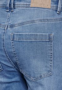 Vaaleansiniset farkut, joissa on sileä rakenne. Niissä on yksi takatasku ja hillitty ommeltu kuvio vyötärönlähellä.