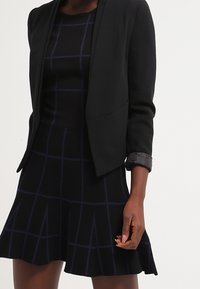 Blazer nero su misura con maniche lunghe. Abbinato a un vestito nero aderente con motivo a griglia blu. Tessuto liscio, design strutturato.