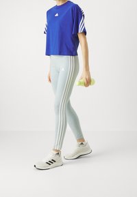 Blå kortärmad t-shirt med vita randar, ljusgröna leggings med tre vita sidostripes och vita sportskor. Håller en grön vattenflaska.