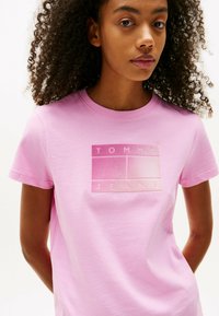 Camiseta de algodón rosa con un parche de logo en relieve y un degradado que presenta el texto "TOMMY JEANS". Mangas cortas y un cuello redondo clásico.