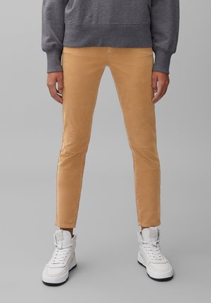 Osoba nosząca dopasowane spodnie w kolorze musztardowym, szarą bluzę i białe wysokie trampki, stojąca na tle gładkiego szarego tła.