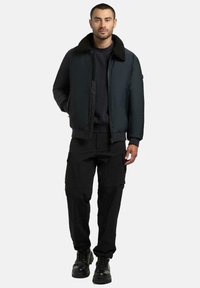 Sort bomberjakke med pelskrage, glidelåslukking og ribbestrikkede mansjetter, kombinert med svarte cargobukser og svarte snøresko.