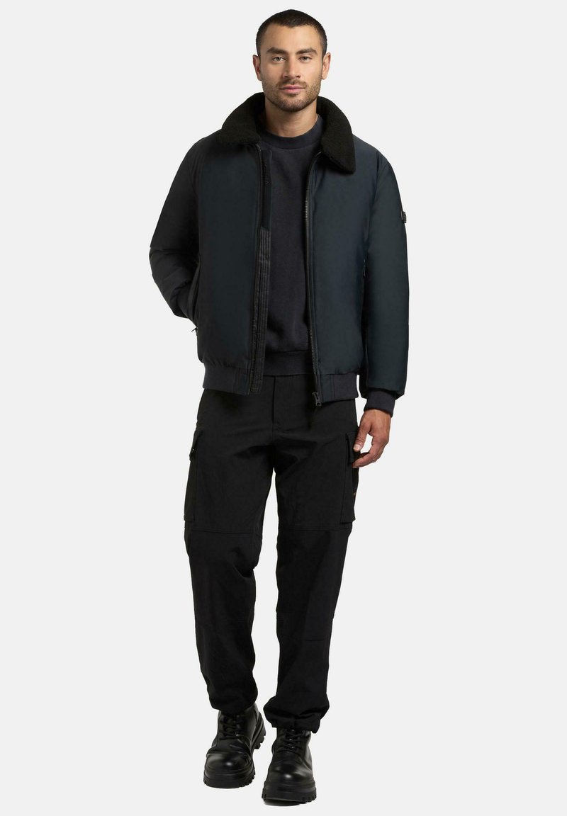 Sort bomberjakke med pelskrage, glidelåslukking og ribbestrikkede mansjetter, kombinert med svarte cargobukser og svarte snøresko.