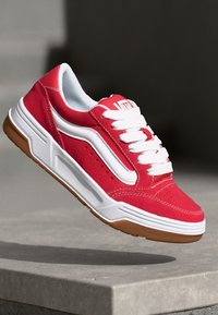 Pantofii sport roșii din suedă, cu șireturi albe, o dungă laterală albă și o talpă din cauciuc texturat de culoare maro. Designul include accente de cusături detaliate.
