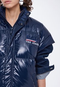 Chaqueta acolchada de color marino con textura brillante, que presenta un cuello alto, cierre de cremallera y un logotipo bordado en el pecho. Detalles de mezclilla en las mangas.