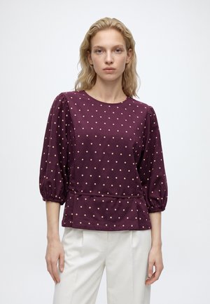 Femme portant un chemisier bordeaux à pois beiges avec des manches bouffantes trois-quarts, associé à un pantalon blanc, debout devant un fond uni.