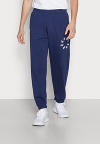 Person klädd i marinblå joggers med vita logotyper på vänster lår, vita sneakers och en vit skjorta, står på en ljusgrå golv.