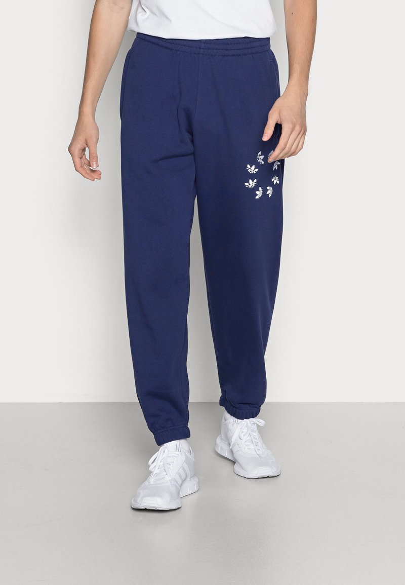 Person klädd i marinblå joggers med vita logotyper på vänster lår, vita sneakers och en vit skjorta, står på en ljusgrå golv.