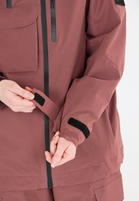 Wasserfester kastanienbrauner Jacke mit schwarzen Reißverschlüssen und Klettverschlüssen an den Ärmeln. Verfügt über eine Fronttasche und ist auf Komfort und Funktionalität ausgelegt.