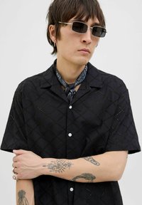 Schwarzes Kurzarmhemd mit Diamantschnittmuster, aus leichtem Stoff. Mit Kragen, durchgehender Knopfleiste und weißen Knöpfen.