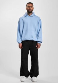 Lys blå oversized hettegenser med lomme foran, kombinert med svarte vide bukser og hvite joggesko. Enkel design og mykt materiale.