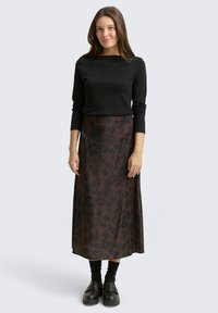 Une femme se tient debout, portant un haut à manches longues noir et une jupe midi à imprimé léopard brun, avec des chaussettes noires et des chaussures noires épaisses.