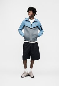 Μπουφάν Nike σε μπλε και γκρι, με φερμουάρ και κουκούλα, συνδυασμένο με μαύρα σορτς. Τα αθλητικά παπούτσια είναι ανοιχτό μπεζ με υφασμάτινες λεπτομέρειες.