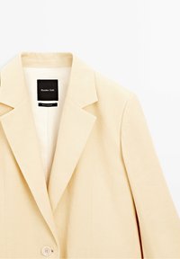 Beżowa, dopasowana marynarka wykonana z gładkiej tkaniny, z wyciętymi klapami, zapinana na jeden guzik, z minimalistyczną metką wewnątrz.