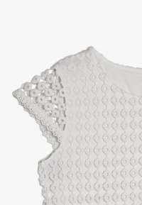 Vit broderad topp med ett geometriskt mönster, texturerad tyg och dekorerad spetskant på axlarna. Rund halsringning.