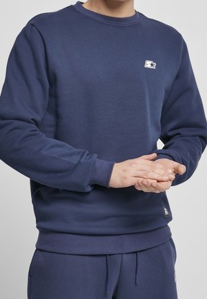 Homme portant un sweat-shirt bleu marine et un pantalon assorti, mains jointes devant, fond clair uni.
