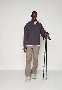 Vyras, vilkintis pilką švarką ir smėlio spalvos kelnes, stovi su dviem juodais žygio lazdomis prieš neutralų šviesų foną.