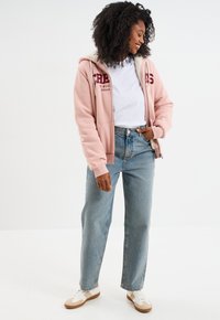 Felpa rosa con fodera bianca, caratterizzata da scritte color bordeaux. Indossata sopra una T-shirt bianca, abbinata a jeans blu chiaro a gamba dritta e sneakers beige.