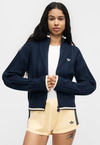 Femme aux longs cheveux foncés porte un pull zippé bleu marine sur un haut blanc et un short jaune clair, avec un tatouage visible sur la cuisse gauche.