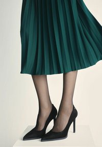 Czarne zamszowe szpilki z ostro zakończonymi noskami i smukłymi obcasami stiletto. Noszone z przezroczystymi czarnymi rajstopami, w zestawieniu z plisowaną zieloną spódnicą.