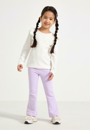 Ung pige med fletninger, iført en hvid langærmet top med flæser, lyse lilla bukser og hvide sneakers, stående foran en ensfarvet baggrund.