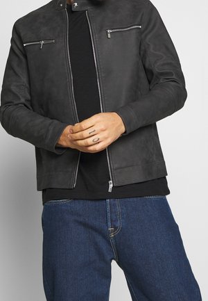 Mann trägt schwarze Lederjacke mit Reißverschluss, schwarzes Hemd und blaue Jeans, mit der Tätowierung "10" auf den Fingern, Hände nahe der Taille gefaltet.