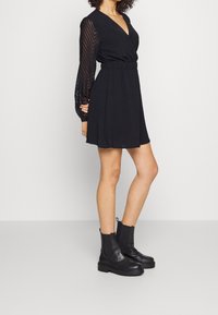 Femme portant une robe noire avec des manches longues en voile à motif et des bottes noires à talons chunky, se tenant devant un fond blanc uni.