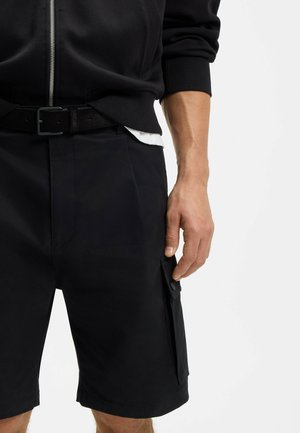 Torso e gambe maschili con pantaloncini cargo neri con tasca laterale e manica di giacca nera con zip tirata fino al gomito.