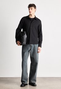 Must musta särk tekstuuriga mustaga nupudega, laiad heledad sinised teksad ja must nahkne käepaelaga kott, hoides ühes käes.