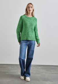 Grün strukturierter Strickpullover mit Rundhalsausschnitt, lockerer Passform und geripptem Saum, kombiniert mit blauen weit geschnittenen Jeans und weißen Sneakern.