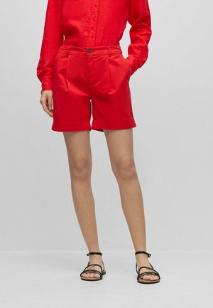 Femme portant une chemise rouge boutonnée et un short rouge, debout les mains dans les poches, sandales noires à brides aux pieds.