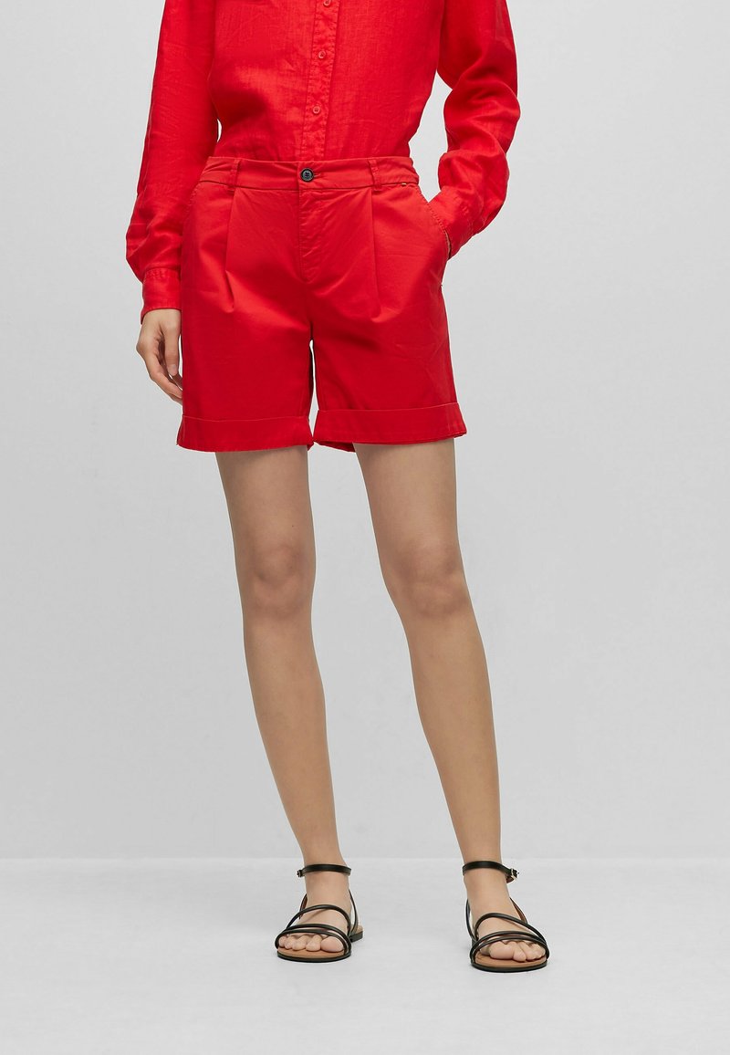 Femme portant une chemise rouge boutonnée et un short rouge, debout les mains dans les poches, sandales noires à brides aux pieds.