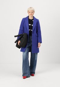 Purple textured coat with a tailored design, worn over a black sweater featuring white heart patterns, paired with wide-leg denim jeans and red heels.