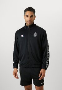 Jachetă de trening neagră cu fermoar, guler înalt, dungi laterale albe și detalii cu logo. Purtați peste pantaloni scurți negri, dintr-un material neted.