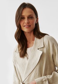 Beige linen trench coat with a belted waist, wide lapels, and long sleeves. Smooth texture with visible stitching details.