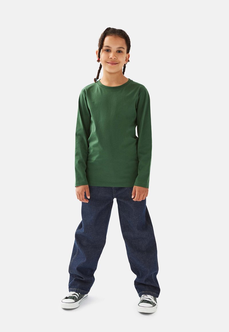 Lächelndes Kind mit geflochtenem Haar steht nach vorne gerichtet, trägt ein grünes Langarmshirt, blaue Jeans und schwarz-weiße Turnschuhe.