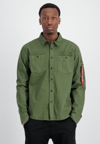 Camisa verde oliva de botones, fabricada en algodón, con dos bolsillos en el pecho, botones negros y una etiqueta de tela roja en la manga izquierda que dice "FLIGHT".
