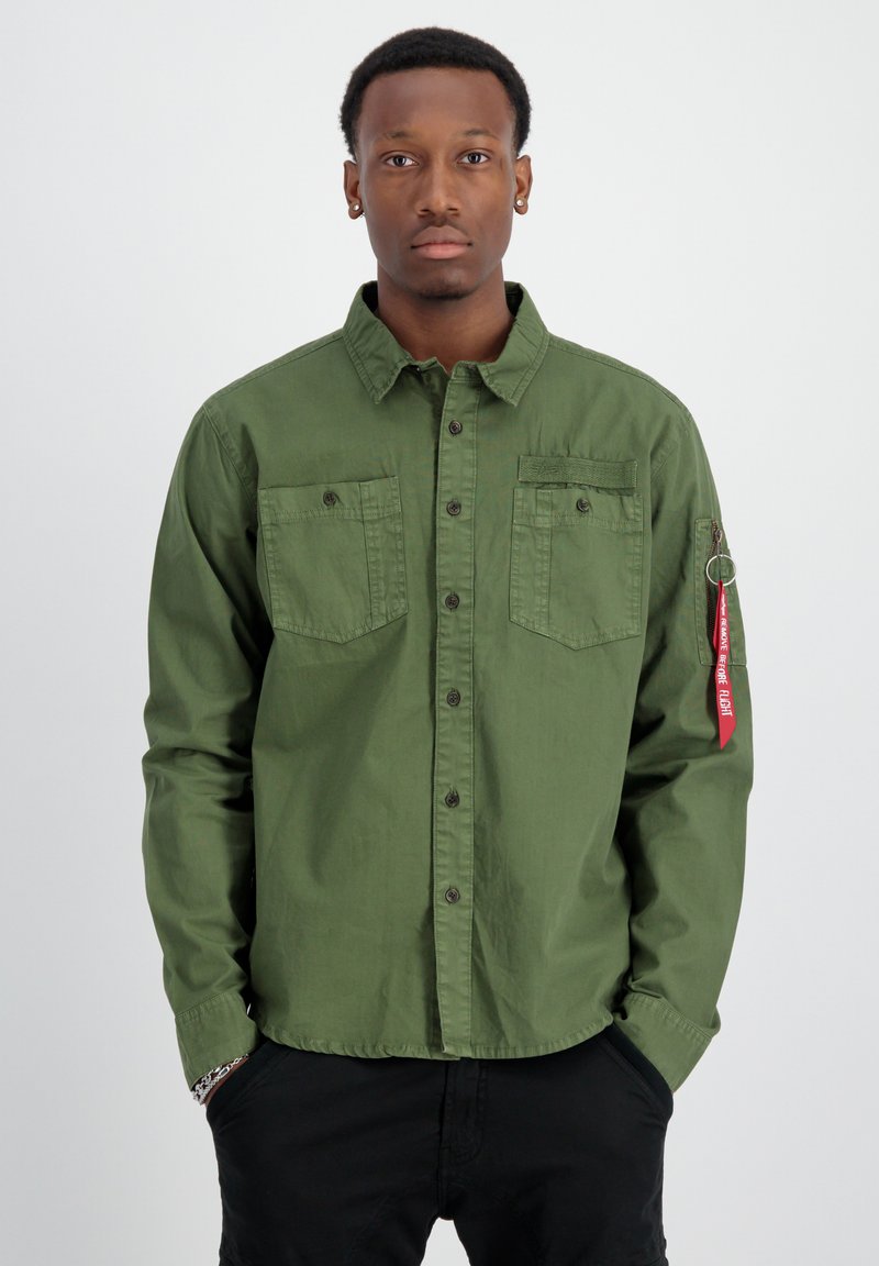 Camisa verde oliva de botones, fabricada en algodón, con dos bolsillos en el pecho, botones negros y una etiqueta de tela roja en la manga izquierda que dice "FLIGHT".