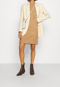 Beige Blazer über einem braunen Strickkleid, accessorisiert mit einer kleinen hellen Handtasche und braunen strukturierten Stiefeletten. Glatte und enganliegende Materialien.