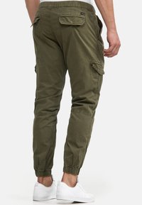 Pantalon cargo vert olive, en tissu doux. Comprend une ceinture élastique, des jambes fuselées et plusieurs poches. Porté avec des baskets blanches.