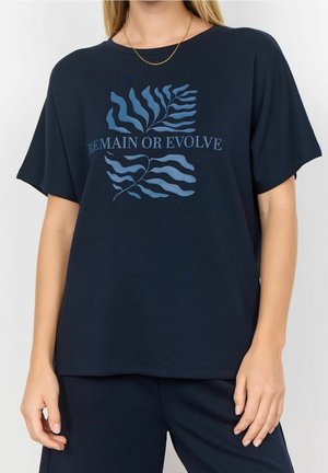 Vrouw draagt een marineblauw T-shirt met een blauw abstract bladontwerp en de tekst "REMAIN OR EVOLVE", gecombineerd met bijpassende marineblauwe broek.