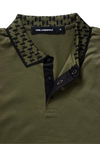 Olivově zelené polo tričko s černým vzorovaným límcem, třemi knoflíky a textilní štítkem. Límec zdobí kontrastní černé detaily.