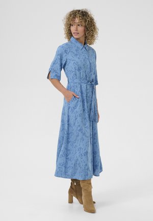 Rochie cămașă de culoare bleumarin deschis, cu model, guler, mâneci scurte, talie legată, buzunare și o siluetă fluidă, asortată cu botine maro cu toc.