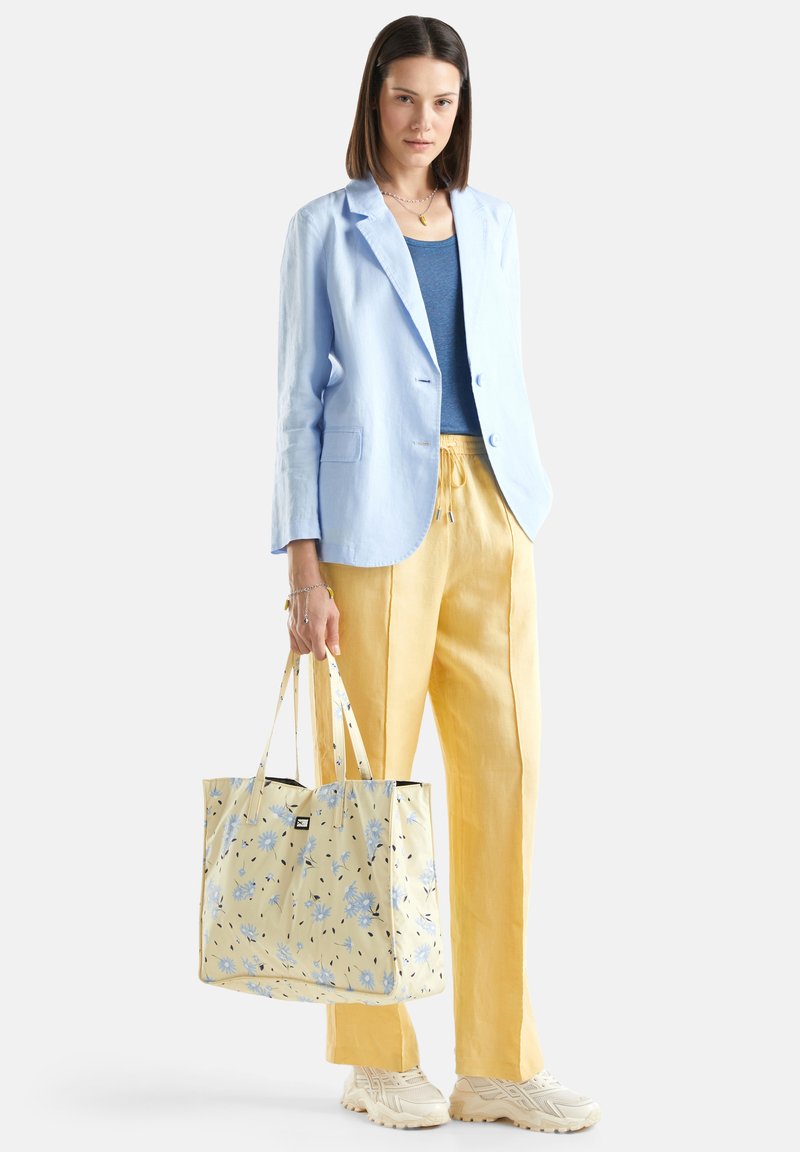 Blazer di lino azzurro chiaro sopra una canotta scura, abbinato a pantaloni di lino gialli. Una borsa a tracolla floreale è tenuta in mano, mentre delle sneakers neutre completano il look.