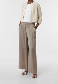Cardigan crème en maille avec manches trois-quarts, porté sur un haut blanc. Pantalon ample de couleur taupe clair avec taille élastique, assorti à des sandales blanches à lanières.