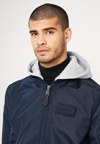 Chaqueta bomber navy con cremallera frontal, capucha gris y cuello acanalado negro. Presenta un parche de logo texturizado y una tela suave y brillante.